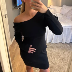 Svart offshoulder kort klänning - Snygg svart kort klänning med offshoulder och lång ärm. Klänningen har en djup öppen rygg och är figurnära med stretchigt material. Perfekt för dig som vill ha en stilren och trendig look.