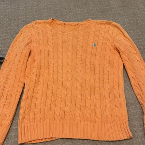 Orange kabelstickad tröja från Polo Ralph Lauren -  Aldrig använt men testat den på .Säljer en orange kabelstickad tröja från Polo Ralph Lauren med klassisk blå logga broderad på bröstet. Tröjan har rund halsringning, ribbade muddar och lång ärm. Perfekt för dig som gillar preppy och färgglada vibes. Lappen klippte vi bort för den kliade. 
