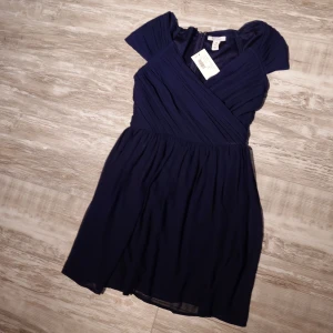 Mörkblå kort klänning från NLY One (oanvänd) - Supersnygg mörkblå kort klänning från NLY One med omlottliv och lätt rynkade cap sleeves. Kjolen är luftig och faller fint, perfekt för dig som gillar en feminin och stilren look. Dragkedja bak för enkel påklädning.