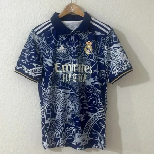 Fotbollströja  - Snygg  fotbollströja i marinblått med unikt drakmönster i vitt. Tröjan har korta ärmar, klassisk krage och guldiga detaljer. Tillverkad i lätt och ventilerande material, perfekt för match eller träning. Officiell produkt med klubbmärke och sponsortryck.