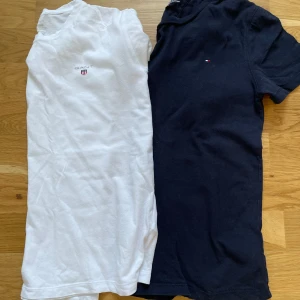 2-pack t-shirts GANT & Tommy Hilfiger - Två klassiska t-shirts: en vit från GANT med liten logga på bröstet och en marinblå från Tommy Hilfiger med diskret flagga. Båda har rund hals, kort ärm och är gjorda i mjuk bomull. Perfekta för en clean och stilren look. Storlek 152