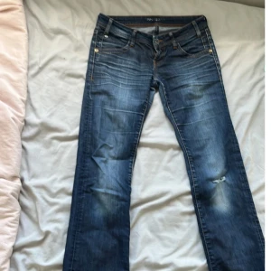 Blå bootcut jeans från Mavi - Snygga blå jeans från Mavi med bootcut passform och klassisk femficksdesign. Jeansen har slitningar och ljusare partier på låren och knäet för en cool, använd look. Tillverkade i mjukt bomullstyg med dragkedjegylf och detaljer i kontrastfärg.