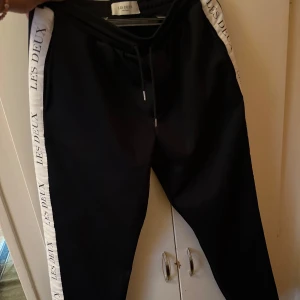 Svarta mjukisbyxor Les Deux med vita revärer - Svarta mjukisbyxor från Les Deux med vita revärer längs sidorna där det står 'LES DEUX' tryckt. Byxorna har snörning i midjan och en bakficka. Perfekta för en chill och sportig look.