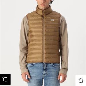 Brun dunväst från Lacoste - Snygg brun dunväst från Lacoste med quiltad design och dragkedja framtill. Västen har en liten grön krokodillogga på bröstet och står ut med sin stilrena look. Perfekt att slänga över en hoodie eller långärmad tröja för extra värme. Köpt för några veckor sedan och knappt använd.