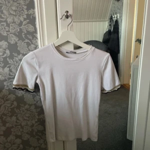 Vit T-shirt med spetsdetalj på ärmarna - Säljer en vit T-shirt med svart, vit och guld detaljer längst ut på ärmarna. Finns inga fläckar eller skador på den, är använd ett få antal gånger. ❤️