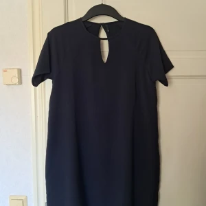 Svart/Mörkblå kortklänning från ONLY - En stilren svart/mörkblå kortklänning från ONLY i storlek 36. Klänningen har korta ärmar, rund halsringning med en snygg öppning fram och knäppning i nacken. Materialet är lätt strukturerat och ger en clean look.