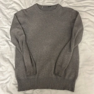 Grå stickad cashmere tröja med rund hals - En stilren grå stickad tröja med rund halsringning och ribbade muddar vid ärmslut och nederkant. Perfekt för lager-på-lager och enkel att matcha med jeans eller chinos. Långärmad och i klassisk modell som funkar året runt. OBS har ett litet hål på vänstra armen.