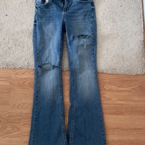Blå bootcut jeans med slitningar - Säljer ett par blå bootcut jeans med snygga slitningar över knäna. Jeansen har låg midja och klassisk femficksdesign. Perfekta för dig som gillar en avslappnad och trendig look. Materialet är jeans i bomull med lite stretch för skön passform.