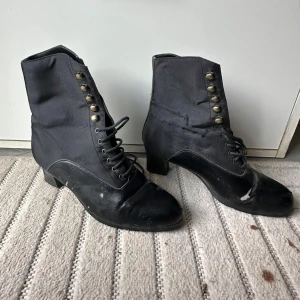 Svarta retro stövletter med snörning - Unika svarta stövletter med snörning och dekorativa knappar i mässing. Skorna har låg klack, rund tå och är tillverkade i en mix av textil och skinn. Perfekta för dig som gillar vintage vibes och vill sticka ut med en klassisk look.