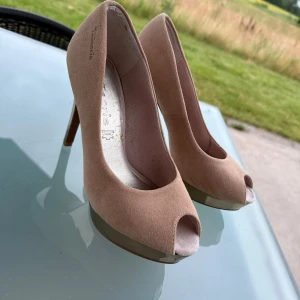 Beige mocka pumps med platå från Tamaris - Snygga pumps från Tamaris i beige mocka med hög klack och platåsula i metallic. Skorna har öppen tå och rundad form framtill. Perfekta för dig som vill sticka ut med en elegant och modern look.
