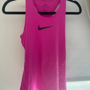 Rosa Nike Dri-Fit linnetopp - Sportig rosa linnetopp från Nike med svart logga på bröstet. Tillverkad i lätt och ventilerande Dri-Fit polyester som håller dig sval under träningen. Ärmlös modell med brottarrygg för maximal rörelsefrihet. Perfekt för dig som gillar att träna med stil. Linnet är i barnstorlek L/146-156! 🤸‍♀️🤽‍♀️