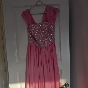 Rosa långklänning med paljetter - Långklänning i rosa med glittrig paljettöverdel och vackert fallande kjol. Klänningen har breda axelband och en romantisk look, perfekt för bal eller fest. Överdelen är dekorerad med silverfärgade paljetter som ger extra glamour.