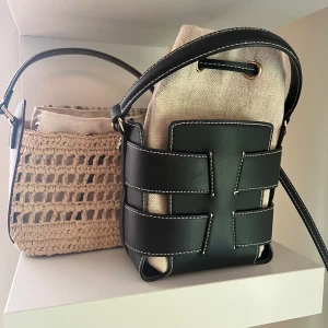 Beige och svart axelväska i canvas och skinn - Snygg axelväska i beige canvas med svarta detaljer i skinn och vita sömmar. Väskan har ett unikt flätat mönster framtill och dragsko upptill. Perfekt för dig som vill ha en trendig och praktisk väska med plats för det viktigaste.