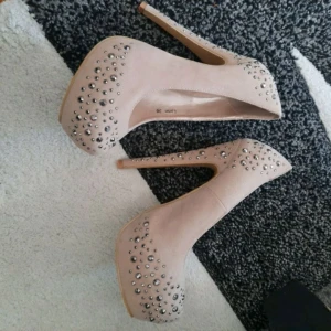 Beige pumps med nitar från NLY - Snygga beige pumps från NLY med höga klackar och plattform. Skorna är dekorerade med silvriga nitar över hela skon och har en rund tå. Ytan är i mockaliknande material som ger en lyxig känsla. Perfekta för dig som vill sticka ut.