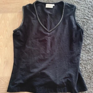 Svart ärmlös linnetopp från Hennes - En enkel svart ärmlös linnetopp från Hennes med v-ringad halsringning och diskreta sömmar längs kanterna. Perfekt att matcha med jeans eller kjol för en clean look. Känns som en M i storleken, egentligen L. Hennes vintage kollektion.