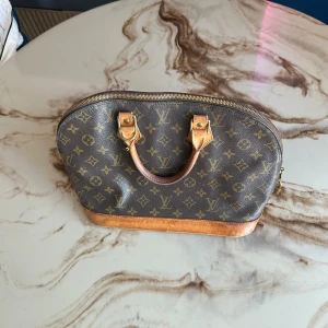 Louis Vuitton Monogram handväska - Klassisk Louis Vuitton handväska i brunt monogrammönster med detaljer i läder och mässingsfärgad dragkedja. Väskan har rundad form, dubbla handtag och ett rymligt innerfack. Ikoniskt LV-lås och snygga kontrastsömmar. Perfekt för dig som gillar lyxig och tidlös stil.