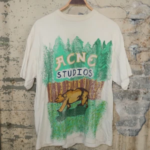 Vit Acne Studios t-shirt med björntryck - Vit t-shirt från Acne Studios med färgglatt tryck av björn, skog och logga på framsidan. Oversized passform och klassisk rund hals. Perfekt statement-plagg för dig som gillar unika prints och streetstyle.