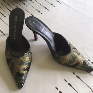 Camo-glittriga klackskor från Rebeca Sanver - Unika pumps med spetsig tå från Rebeca Sanver, täckta i glittriga paljetter i camouflage-mönster. Skorna har hög klack, öppen häl och svart innersula. Perfekta för dig som vill sticka ut med en edgy och glam look.