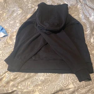 Svart hoodie med magficka - En klassisk svart hoodie med huva och stor magficka framtill. Tröjan har långa ärmar och ribbade muddar vid ärmslut och nederkant. Perfekt för chill dagar eller när du vill ha en enkel och stilren look.