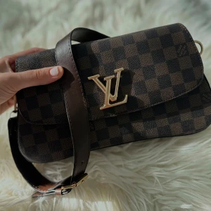 Louis Vuitto rutig axelväska brun - Säljer en snygg axelväska från Louis Vuitto i klassiskt brunt rutigt mönster med guldigt spänne. Väskan har en bred axelrem och är gjord i skinn. Perfekt storlek för det viktigaste och ger en lyxig touch till din outfit.