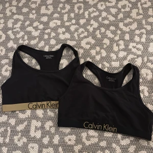 Svarta bralette från Calvin Klein 2-pack - Två svarta braletter från Calvin Klein med bred resår och guldigt logoband. Sportig modell med brottarrygg och stretchigt material som sitter skönt. Perfekt för dig som gillar enkel och stilren design med tydlig logga.