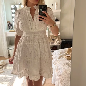 Vit kort klänning med spetsdetaljer - Superfin vit kort klänning i bomull med broderade spetsdetaljer och volangkant. Klänningen har v-ringning med små knappar, puffiga korta ärmar och markerad midja. Perfekt för sommarens alla tillfällen och ger en romantisk vibe. Har lite sminkfläckar men borde gå bort enkelt!