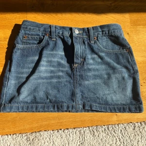 Blå jeanskjol från Unlimited - Snygg blå jeanskjol från Unlimited med klassisk femficksdesign, bälteshällor och knappstängning fram. Kjolen är kort och har coola bakfickor med en liten röd detalj på ena sidan. Jättebra skick!