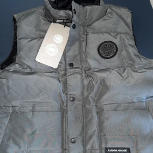 Grå dunväst från Canada Goose - Snygg grå dunväst från Canada Goose med svart logotypmärke på bröstet och två stora fickor framtill. Västen har hög krage, tryckknappar och är quiltad för extra värme. Perfekt för lager-på-lager och streetstyle.