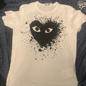 Vit t-shirt Comme des Garçons Play - Vit t-shirt från Comme des Garçons Play med stort svart hjärta och ögon på bröstet, omgiven av coola svarta färgstänk. Klassisk passform och rund hals. Perfekt statement-plagg för dig som gillar streetstyle och unika prints. Skick 9/10. Vid snabbt köp kan pris diskuteras 