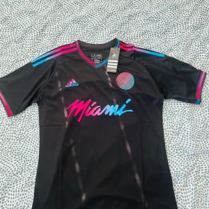Inter Miami CF svart matchtröja Adidas - Snygg svart Inter Miami CF fotbollströja från Adidas med korta ärmar. Tröjan har rosa och blå detaljer på axlar och ärmslut, samt klubbmärke och Adidas-logga på bröstet. Miami-tryck i rosa och blått framtill. Tillverkad i lätt och ventilerande polyester.
