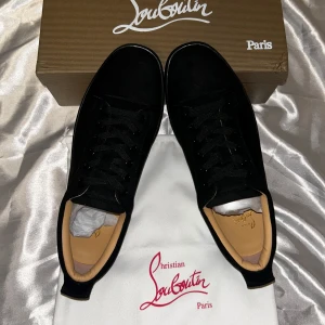 Svarta sneakers från Christian Louboutin - Snygga svarta sneakers från Christian Louboutin i mocka med klassisk röd sula och diskret broderad logga på hälen. Skorna har svarta snören, rund tå och beige innersula med gulddetalj. Perfekta för dig som vill ha en lyxig och stilren look.