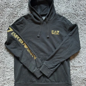 Svart EA7 Emporio Armani hoodie - Svart hoodie från EA7 Emporio Armani med gul logga på bröstet och stort tryck längs ena ärmen. Klassisk huva med snörning och känguruficka framtill. Skön passform och perfekt för en chill och stilren look.