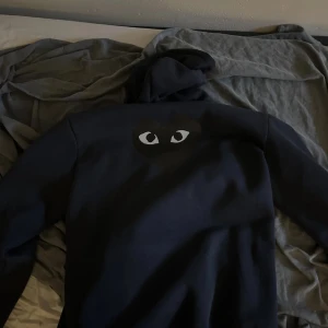 Mörkblå hoodie Comme des Garçons Play - Säljer en svart hoodie från Comme des Garçons Play med det ikoniska hjärtat med ögon på bröstet. Tröjan har huva med snörning, dragkedja framtill och två fickor. Perfekt för dig som gillar streetwear och vill ha något stilrent men ändå unikt.