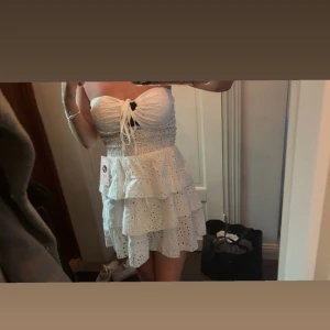 Vit axelbandslös volangklänning - Supersöt vit kortklänning med volanger och broderade hål, axelbandslös modell med smock i midjan och knyt framtill. Perfekt för sommarens varma dagar och har en romantisk vibe.