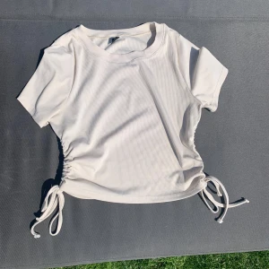 Ljus beige ribbad topp med snörning i sidan - Superfin ljus beige ribbad topp med kort ärm och snörning i båda sidorna som ger en snygg croppad look. Perfekt till jeans eller kjol för en fräsch och enkel stil. Mjuk och stretchig, passar perfekt till varma dagar.