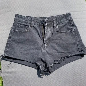 Svarta jeansshorts - Snygga svarta jeansshorts med hög midja och rå, fransig kant nertill. Klassisk femficksmodell med dragkedja och knapp framtill. Perfekta för sommaren och ger en cool, avslappnad vibe.