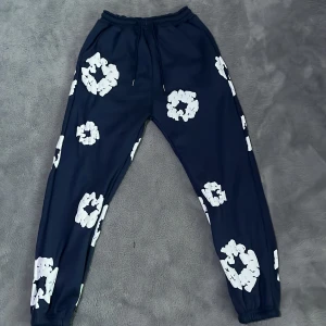 Mörkblå mjukisbyxor Denim Tears - Säljer ett par helt nya mörkblå mjukisbyxor från Denim Tears med vita blommiga tryck över hela byxorna. Byxorna har resår och snörning i midjan samt muddar vid bensluten. Perfekta för en chill och trendig look.