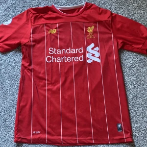 Liverpool FC matchtröja M.Salah - Liverpool FCs officiella matchtröja i rött med tunna vita ränder, broderat klubbmärke och sponsorloggor. Korta ärmar, Premier League-märke på ena ärmen och Western Union-tryck på den andra. Tillverkad i lätt och ventilerande funktionsmaterial.