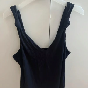 Svart linne med knut - Supersnygg svart linne med breda axelband och coola knutdetaljer på axlarna. Klänningen är ribbad och har en enkel, rak passform som känns både trendig och avslappnad. Perfekt för dig som gillar stilrena plagg med en twist