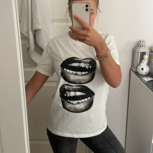 Vit t-shirt med svart läppar-tryck - Vit t-shirt med två stora svarta läppar-tryck framtill. Klassisk rund halsringning och korta ärmar. T-shirten har en normal passform och lite oversized, perfekt för en cool och avslappnad stil.