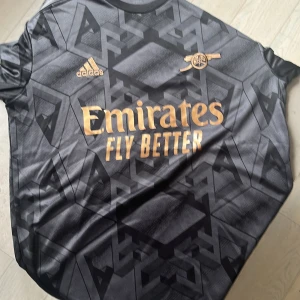 Arsenal bortatröja 22/23 G.Jesus #9 - Snygg Arsenal bortatröja från Adidas med G.Jesus och nummer 9 på ryggen. Tröjan är svart med geometriskt mönster och guldiga detaljer som klubbmärke och sponsortryck. Tillverkad i lätt och ventilerande material, perfekt för match eller träning. Skick 9/10, fotbollströjkollektion bild 5.