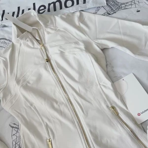 (Nytt Skick) Lululemon Define Jacket i färgen vitt 🤍 - Supersnygg vit jacka från Lululemon med guldiga dragkedjor och figurnära passform. Jackan har hög krage och långa ärmar, perfekt för en sportig och clean look. Materialet känns mjukt och stretchigt, vilket gör den bekväm att bära hela dagen. (Kommer med påse och tag 🏷️ på)