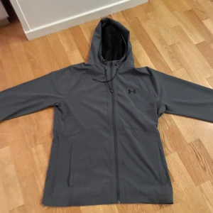 Grå vindjacka från Under Armour - Snygg grå vindjacka från Under Armour med huva och dragkedja framtill. Jackan har minimalistisk design, två fickor och diskret logga på bröstet. Perfekt för blåsiga dagar och outdoor-aktiviteter. Materialet är lätt och vattenavvisande.