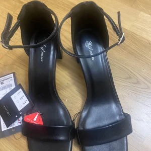 Svarta sandaler med klack från Amisu - Stilrena svarta sandaler från Amisu i storlek 38. Skorna har fyrkantig tå, bred rem över foten och smal ankelrem med spänne. Platt sula med låg klack och tillverkade i syntetmaterial. Perfekta för en trendig look.