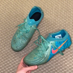 Nike Phantom GX - Snygga turkosa fotbollsskor från Nike med rosa och orange detaljer. Skorna har snörning och en strömlinjeformad design för optimal prestanda på planen. Perfekta för dig som vill sticka ut med stil.