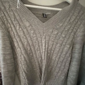 Grå kabelstickad tröja från H&M - Mysig grå kabelstickad tröja från H&M med v-ringning och långa ärmar. Perfekt för lager-på-lager och chill dagar. Tröjan har en klassisk stickad struktur och bred ribbad kant vid halsen.