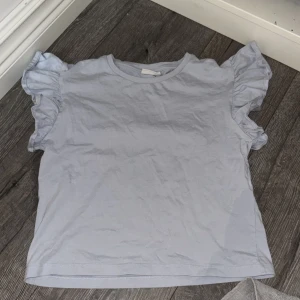 Ljusblå volangtopp från Kappahl - Supersöt ljusblå topp från Kappahl med volangdetaljer på ärmarna. T-shirten har rund halsringning och är tillverkad i mjuk bomull, perfekt för en chill och stilren look. Passar dig som gillar enkla men ändå unika plagg.