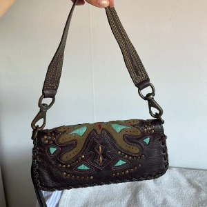 Brun axelväska med broderad fjäril - Unik brun axelväska från Parcel i skinn med broderad fjäril. Väskan har avtagbar axelrem, dekorativa nitar och dragkedja runtom. Insidan har flera kortfack. Kan ta bort axelrem och ha som plånbok. 