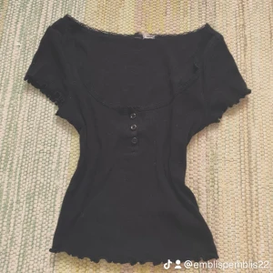 Svart ribbad topp med spets och knappar - Supersöt svart ribbad topp med korta ärmar, dekorativa knappar framtill och spetskant runt den vida urringningen. Ärmsluten har volangdetaljer som ger en gullig touch. Perfekt för dig som gillar en feminin och trendig stil. Från H&M