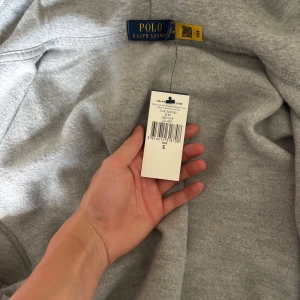 Grå hoodie från Polo Ralph Lauren - Snygg grå hoodie från Polo Ralph Lauren med klassisk broderad logga på bröstet. Tröjan har huva med vita snören, dragkedja framtill och två fickor. Tillverkad i mjuk bomullsmix som är perfekt för chill dagar eller när du vill ha en clean look. Helt ny, då den var för liten för mig, priset går verkligen att tryckas ner vid snabb affär, köpt på zalando